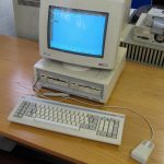 An Amstrad PC1512 with its colour monitor on top of the system unit, a keyboard and the Amstrad mouse, on a desk