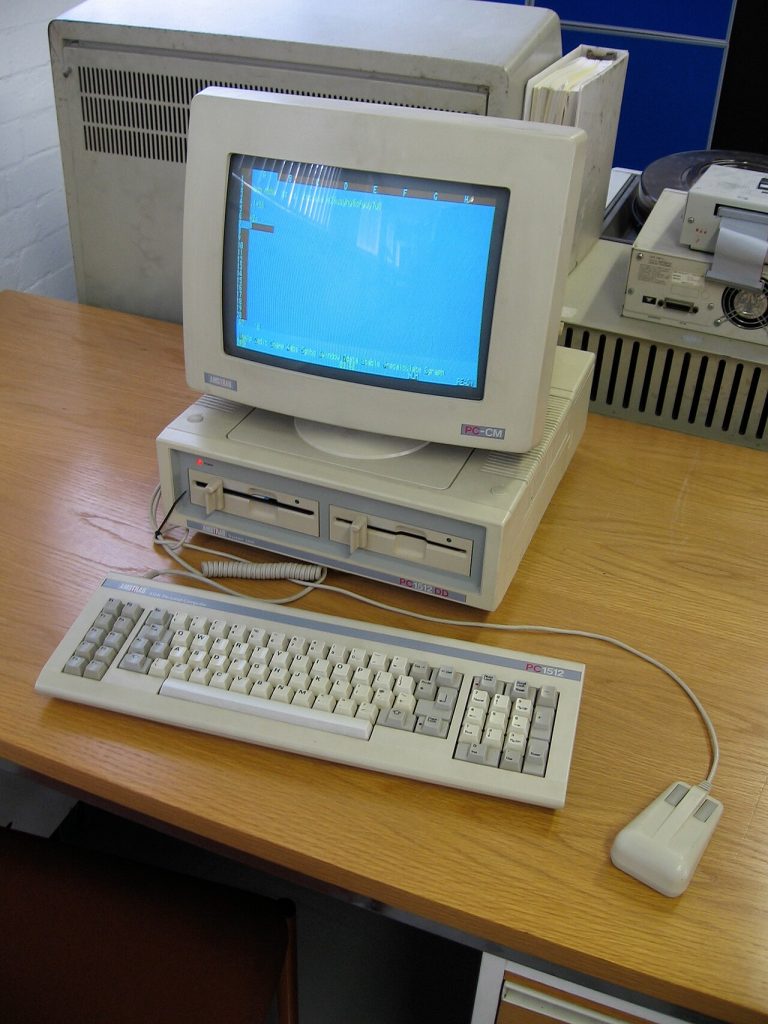An Amstrad PC1512 with its colour monitor on top of the system unit, a keyboard and the Amstrad mouse, on a desk