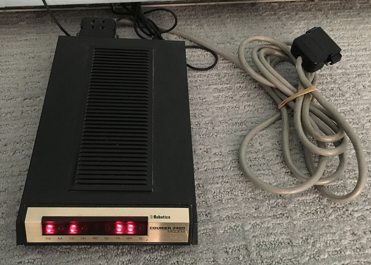 A black USRobotics Courier 2400 external modem with its red status lights on and a grey serial cable