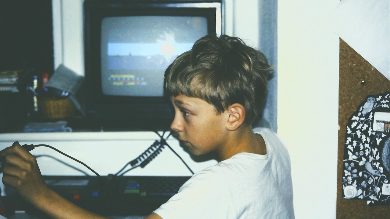 The author as a boy in a white T-shirt at his Amstrad CPC 464, holding the light pen cable, with Beach-Head on the monitor